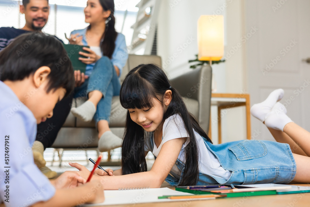 Fototapeta premium Happy Asian family relaxing on sofa while kid drawing on floor. Little boy girl having fun, friendship between siblings, family leisure time in living room at home.