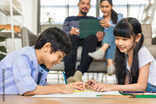 Happy Asian family relaxing on sofa while kid drawing on floor. Little boy girl having fun, friendship between siblings, family leisure time in living room at home.
