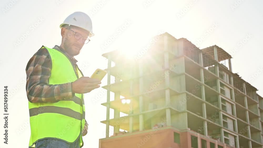 Young male builder engineer architect foreman designer typing on the phone, looking at the project against the backdrop of the construction of a multi-storey building. Slow motion footage Young male builder engineer architect foreman designer typing on the phone, looking at the project against the backdrop of the construction of a multi-storey building. Slow motion footage