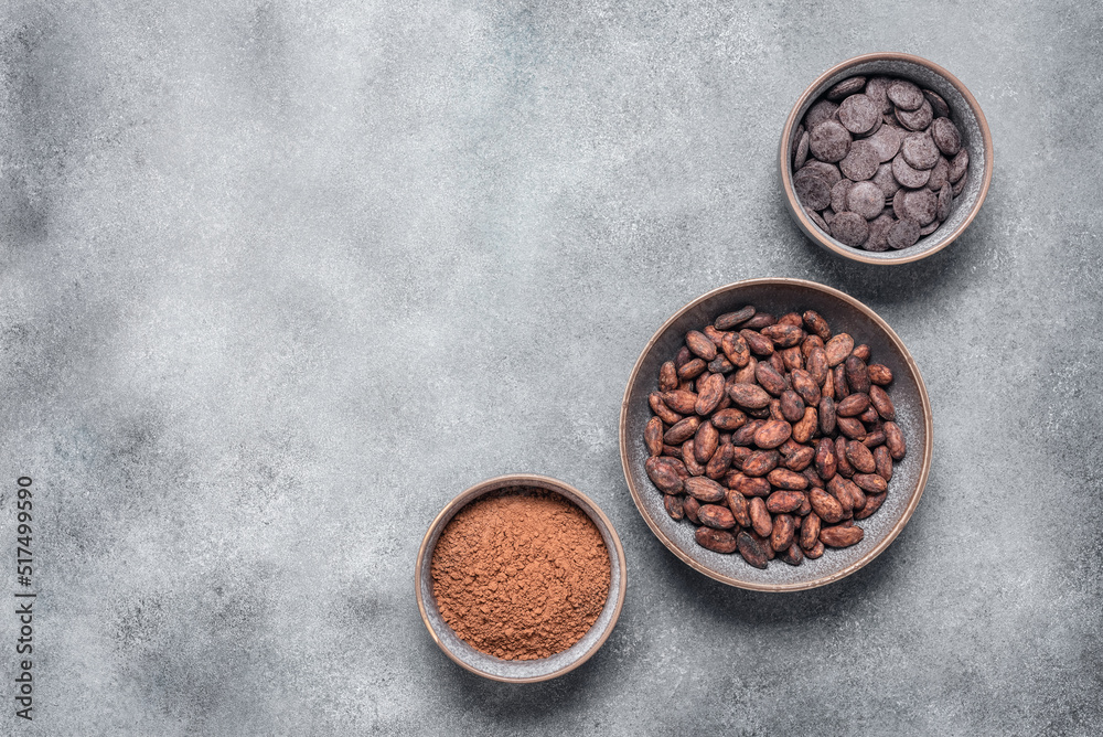 Fototapeta premium Cocoa beans, cocoa powder and chocolate in bowls on grunge gray background. Top view, flat lay.