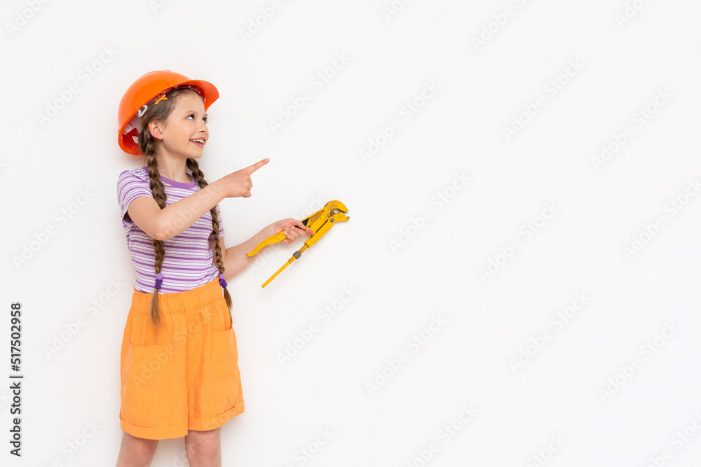 A little girl in an orange construction helmet holds a monkey wrench and points at your advertisement on a white isolated background. Copy space. A little girl in an orange construction helmet holds a monkey wrench and points at your advertisement on a white isolated background. Copy space.
