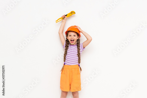 A little girl in an orange construction helmet with two pigtails raises a monkey wrench and smiles broadly on a white isolated background. The concept of renovation in the children's room.