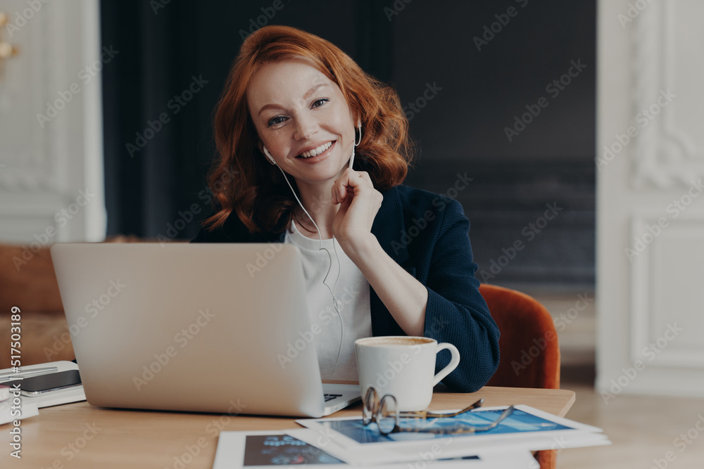 Freelance business woman with ginger hair sits in front of laptop computer, communicates with colleagues via video conference, sits at desktop, drinks coffee, has happy expression. Distance job Freelance business woman with ginger hair sits in front of laptop computer, communicates with colleagues via video conference, sits at desktop, drinks coffee, has happy expression. Distance job