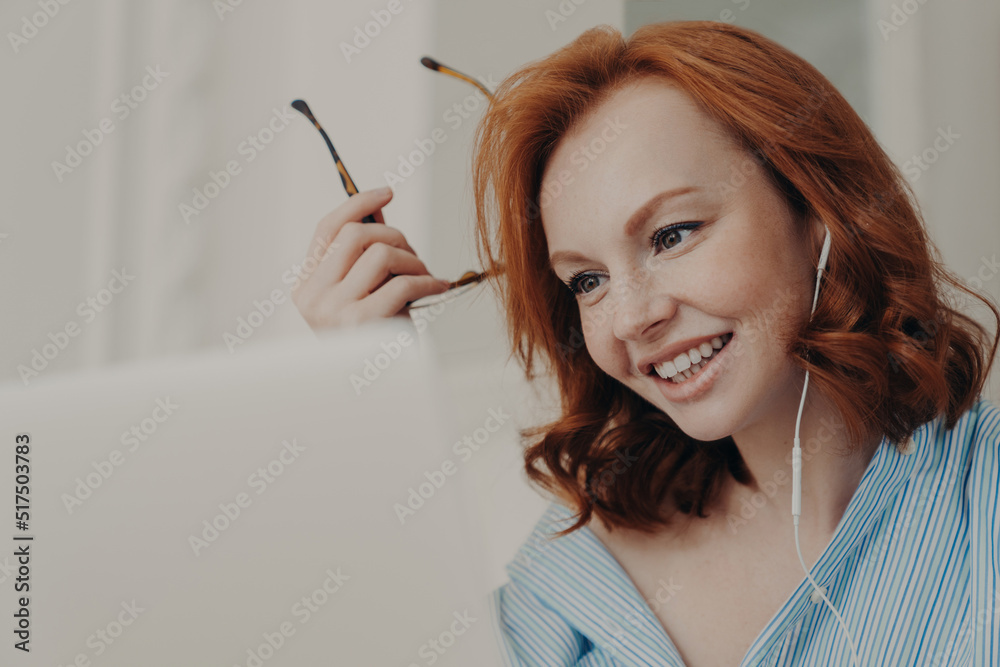 Obraz premium Close up shot of redhead businesswoman makes video call to business partner, has discussion with corporate partner, holds optical glasses, smiles pleasantly, uses earphones. Remote job interview