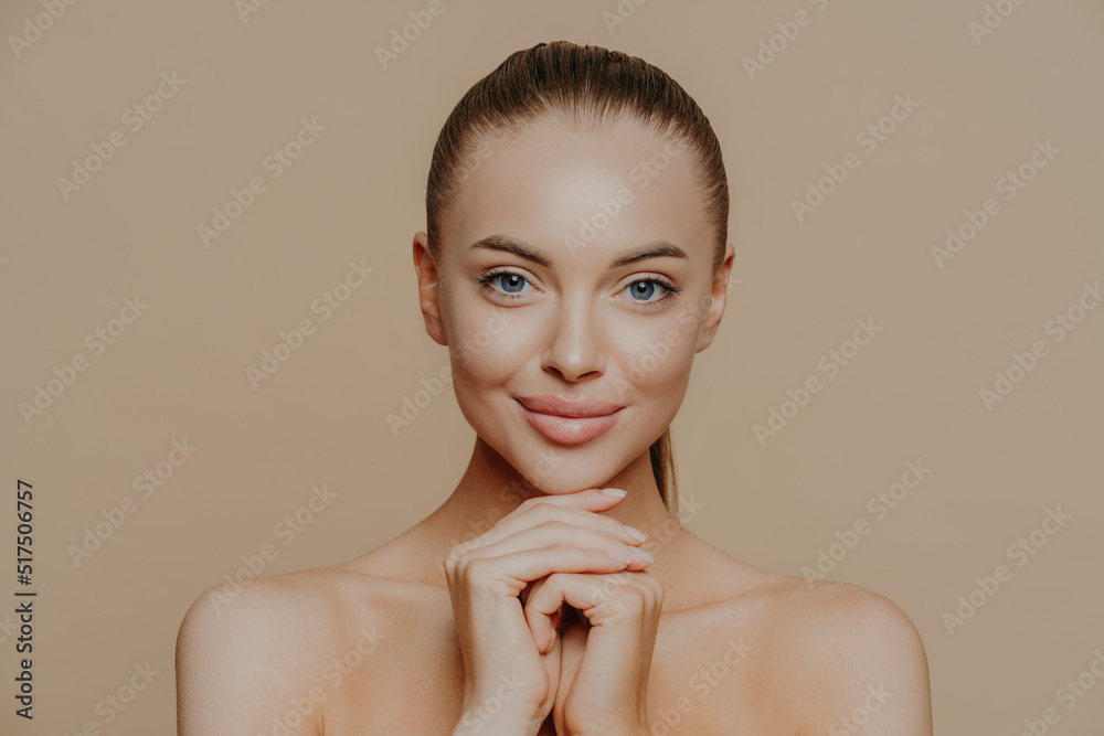 Young pretty woman with beauty skin and combed hair, keeps hands under chin, has healthy complexion after face purification, soft well cared body, isolated over studio wall. Facial treatment Young pretty woman with beauty skin and combed hair, keeps hands under chin, has healthy complexion after face purification, soft well cared body, isolated over studio wall. Facial treatment