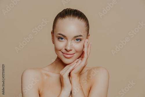 Natural beauty portrait. Photo of young woman enjoys perfect fresh skin after visiting cosmetologist, smiles gently, poses naked against beige background, touches face, wears minimal makeup.