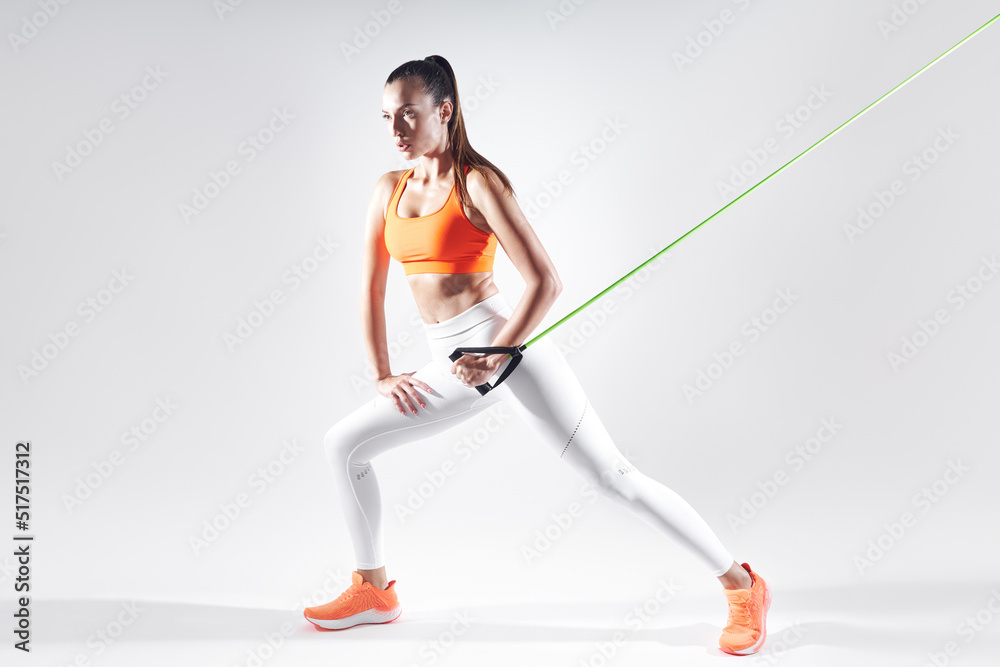 Fototapeta premium Confident woman in sports clothing using resistance band while exercising against white background
