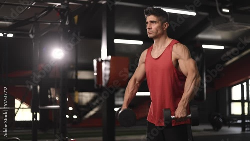 Tough man standing in the gym and pumping his hands with dumbells
