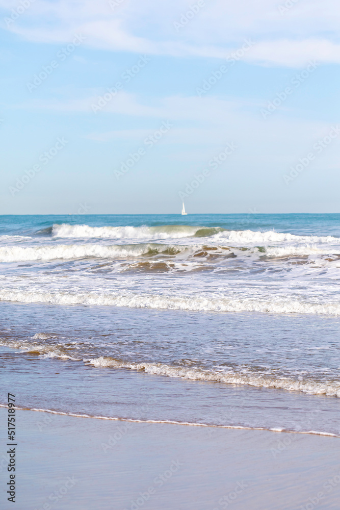 Summer vibes on the sunny autumn beach of Malvarrosa in Valencia, Spain. Sailing yachts in the sea floating along the bubbling foamy waves.