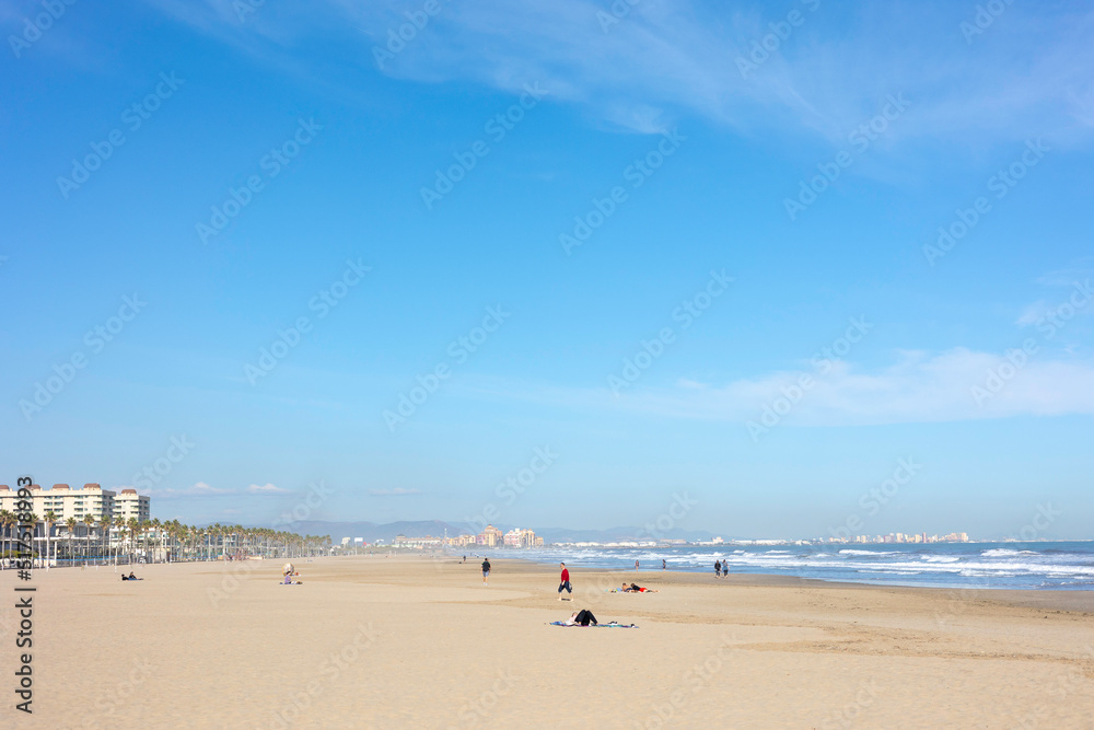 Summer vibes on the sunny autumn beach of Malvarrosa in Valencia, Spain. Vast expanses of smooth fine sand on the sea coast attract vacationers to solitary walks along the bubbling foamy waves.