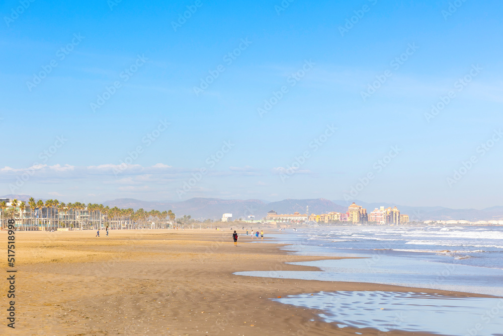 Summer vibes on the sunny autumn beach of Malvarrosa in Valencia, Spain. Vast expanses of smooth fine sand on the sea coast attract vacationers to solitary walks along the bubbling foamy waves.