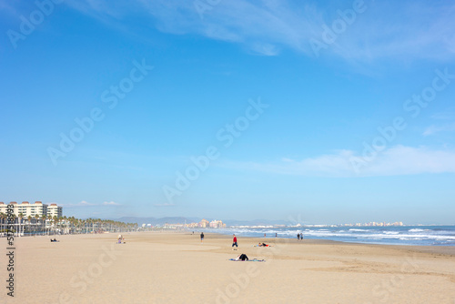 Summer vibes on the sunny autumn beach of Malvarrosa in Valencia, Spain. Vast expanses of smooth fine sand on the sea coast attract vacationers to solitary walks along the bubbling foamy waves. © Sodel Vladyslav