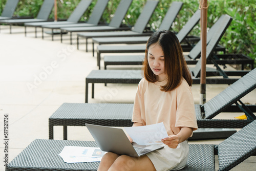 close-up A woman working at the swimming pool with her laptop computer holding paperwork in her hand. It is imperative for young Asian female tourists to sit and work on vacations at the hotel.