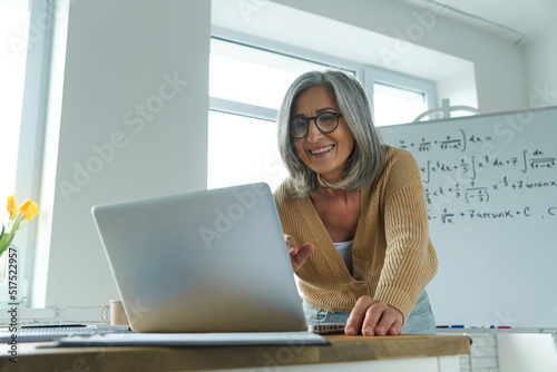 Mature woman teaching mathematics while standing near the whiteboard and looking at laptop