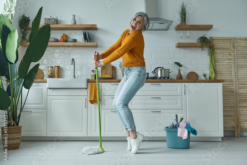 Photo Playful senior woman dancing with a mop while standing at the domestic kitchen