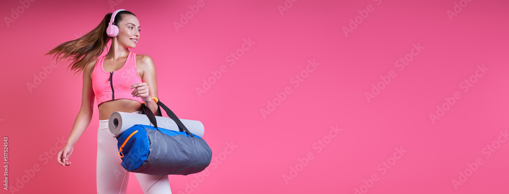 Happy young woman in sports clothing carrying a bag against pink background Happy young woman in sports clothing carrying a bag against pink background