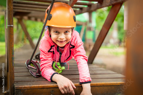 Portrait of little smiling girl in helmet and harness on trail in sky rope park in summer © Olena Shvets