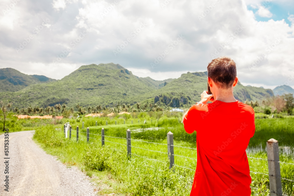 Travel man in Asia trip on Thailand holidays. Traveler blogger in red t-short shooting photo or video of beautiful mountain covered with greenery on mobile phone. Travel and active life concept.