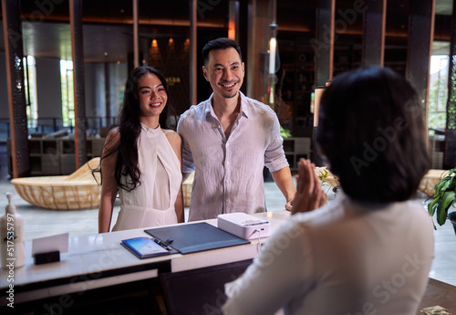 Young couple walking in lobby and arriving at hotel reception while front desk employee welcoming and greeting them