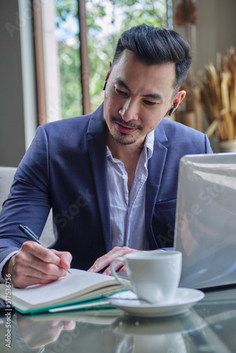 Young handsome asian businessman using laptop and writing notes, sitting in cafe or coworking space. Conference call or working online