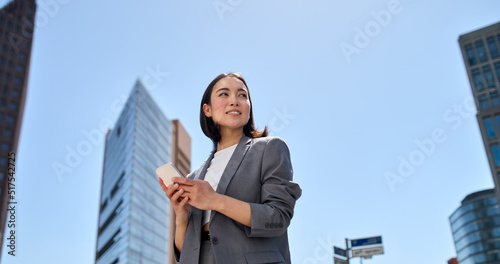 Young busy successful beautiful Asian business woman, korean professional businesswoman holding cellphone using smartphone standing or walking on big city urban street outside.