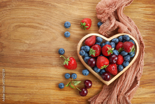 Berries mix in in a heart shaped bowl spilled on rustic wooden table