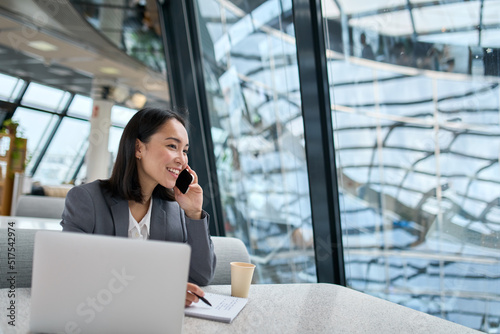 Fotobehang Young happy Asian business woman company employee or sales manager wearing suit working in contemporary glass office talking to client on the phone using laptop working in big modern work space