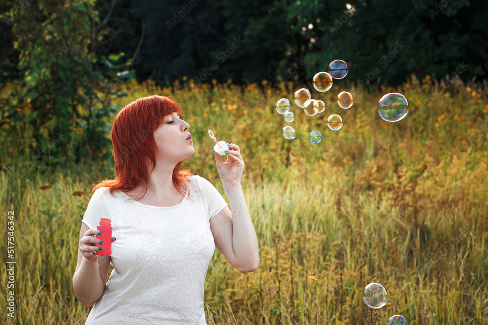 Young red haired woman blowing soap bubbles. Happy girl in nature in the sun.