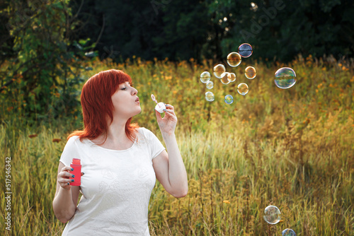 Young red haired woman blowing soap bubbles. Happy girl in nature in the sun.