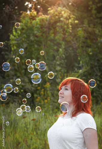 Young woman in a white T-shirt is playing with soap bubbles. Happy red-haired girl in the sun.