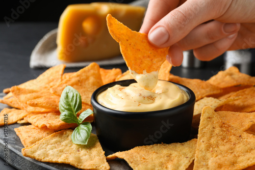 Woman dipping crispy nacho into delicious cheese sauce at table, closeup photo