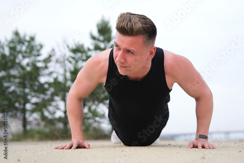Portrait of handsome sportive fit fitness man, athlete, athletic guy doing push ups workout exercise on ground, training outdoors at summer sunny day in park. Healthy lifestyle. Open air gym
