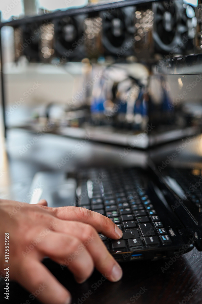 Close up of man's hand while typing on laptop keyboard, cryptocurrency mining rig in background Close up of man's hand while typing on laptop keyboard, cryptocurrency mining rig in background