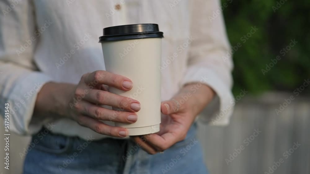 Paper glass of takeaway coffee tea in hands close up. Girl in white linen shirt and blue shorts holds paper coffee cup in the park. Woman is enjoying drink and walk in green park mockup. Slow motion