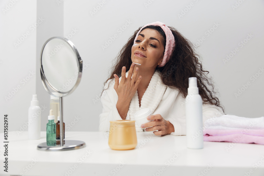 Joyful tanned pretty curly Latin lady apply facial cream at chin admiring herself in mirror doing beauty procedure in home interior. Copy space. Aesthetic medicine skincare beauty products ad concept Joyful tanned pretty curly Latin lady apply facial cream at chin admiring herself in mirror doing beauty procedure in home interior. Copy space. Aesthetic medicine skincare beauty products ad concept