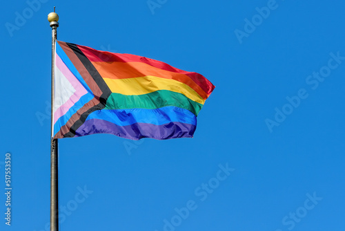 A LGBTQ+ rainbow progress pride flag flaps from a flagpole in a brisk breeze. Blue sky behind.