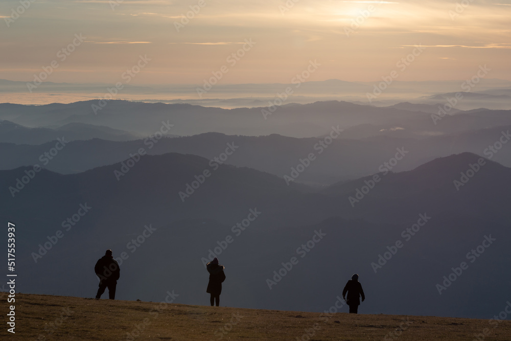 Three people silhouettes on top of a mountain at sunset, looking at endless layers of hills and mountains.