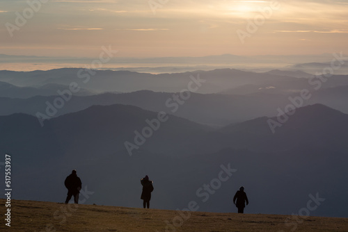 Three people silhouettes on top of a mountain at sunset, looking at endless layers of hills and mountains. © Massimo