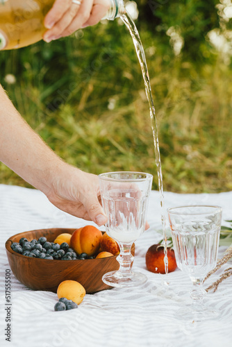 Summer picnic aesthetics. Hands pouring champagne in glasses, tasty fruits and berries in bowl, wildflowers on blanket. Blueberries, peaches and apricots. Vacation and family time concept
