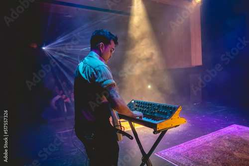 Young latino bassist and keyboardist with shirt and glasses, and cream bass and vintage keyboard playing live in a concert under colorful lights photo