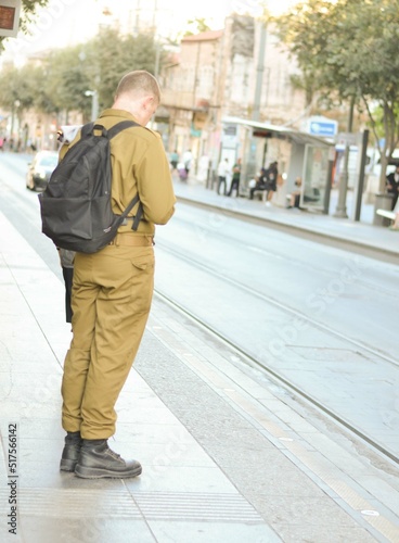 Israeli jewish soldier wearing a green army uniform and black backpack waiting at the train station looking at his phone on his way to base or going home to his family
