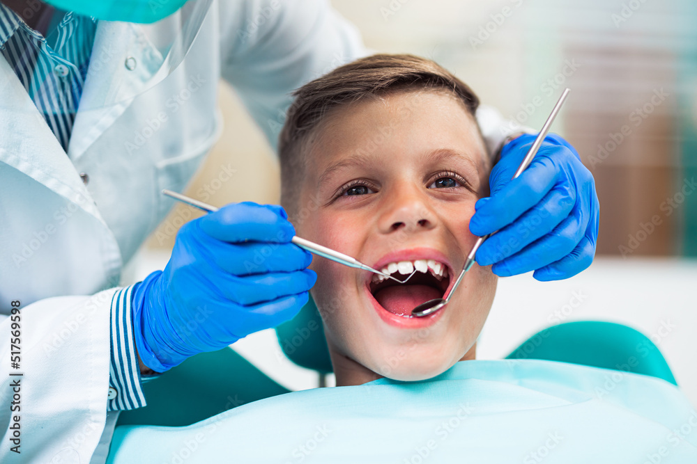 Cute little boy sitting on dental chair and having dental treatment. Dentist is wearing protective face mask due to Coronavirus pandemic. Cute little boy sitting on dental chair and having dental treatment. Dentist is wearing protective face mask due to Coronavirus pandemic.