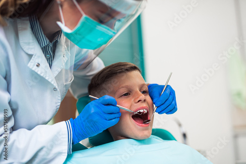 Cute little boy sitting on dental chair and having dental treatment. Dentist is wearing protective face mask due to Coronavirus pandemic.