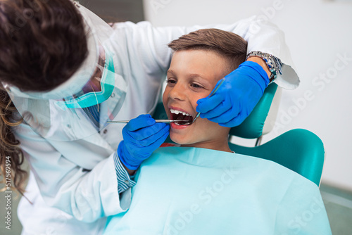 Cute little boy sitting on dental chair and having dental treatment. Dentist is wearing protective face mask due to Coronavirus pandemic.