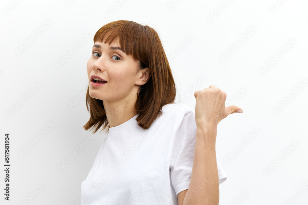 a sweet, emotional, playful woman in a white T-shirt stands on a white background and points with her thumb towards an empty space a sweet, emotional, playful woman in a white T-shirt stands on a white background and points with her thumb towards an empty space
