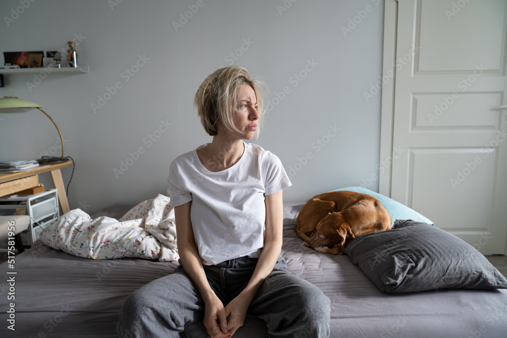 Mental disorders in mid-life. Unhappy depressed middle-aged woman living alone with dog sit on bed looking into distance, thinking about personal problems, feeling lonely, suffering from depression Mental disorders in mid-life. Unhappy depressed middle-aged woman living alone with dog sit on bed looking into distance, thinking about personal problems, feeling lonely, suffering from depression