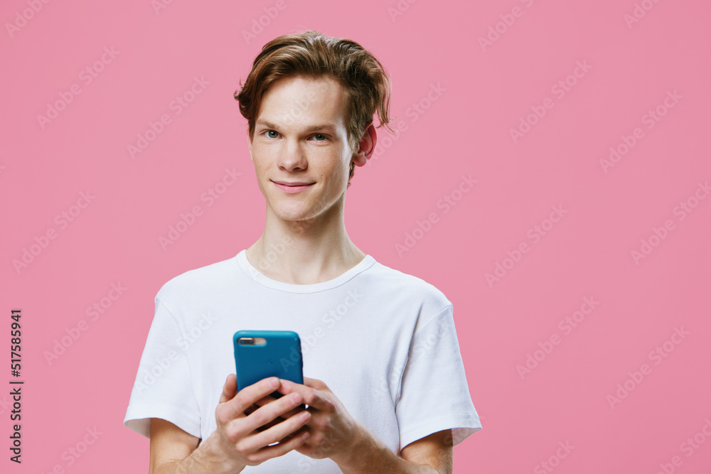 a handsome attractive guy is standing in a white T-shirt and smiling pleasantly at the camera holding the phone in his hand. Horizontal photo on a pink background with space for text a handsome attractive guy is standing in a white T-shirt and smiling pleasantly at the camera holding the phone in his hand. Horizontal photo on a pink background with space for text
