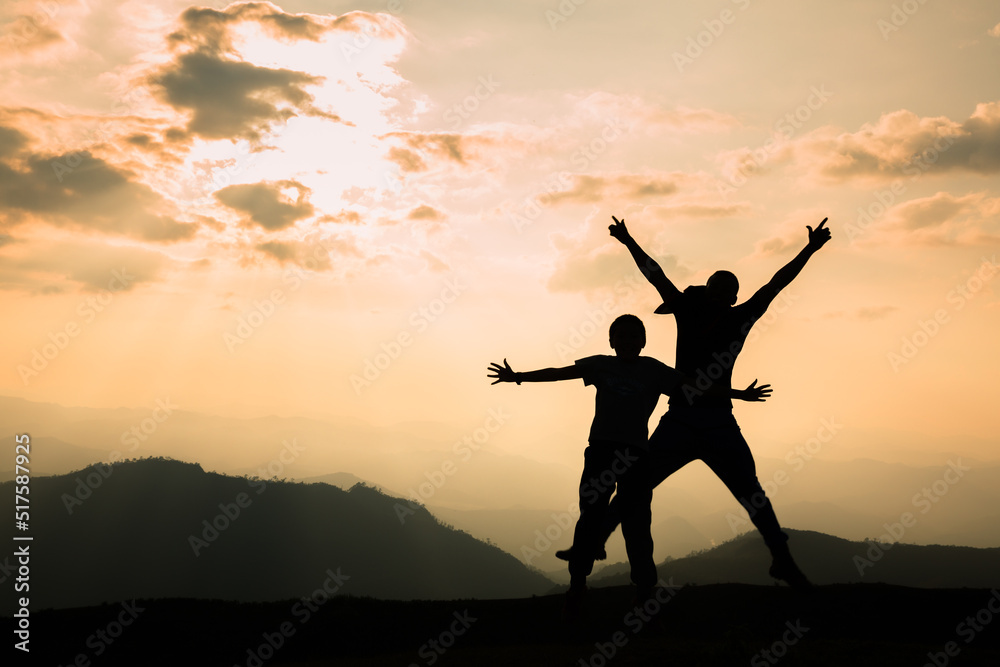 Two climbers in silhouette stand on a beautiful mountain with their hands raised against an orange sky background.