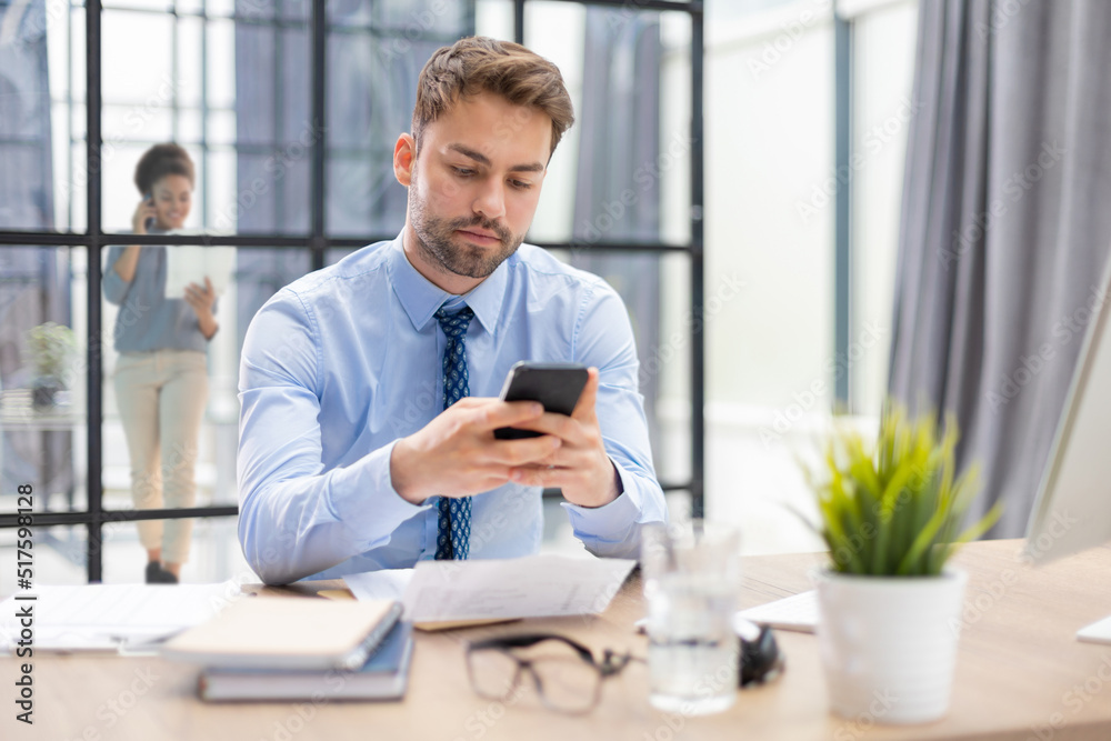 Handsome businessman in office using smartphone with collegues on the background.
