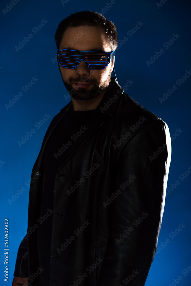 portrait of a man in a studio in a black leather coat. model with a beard on a blue background. trendy luminous glasses on the face portrait of a man in a studio in a black leather coat. model with a beard on a blue background. trendy luminous glasses on the face